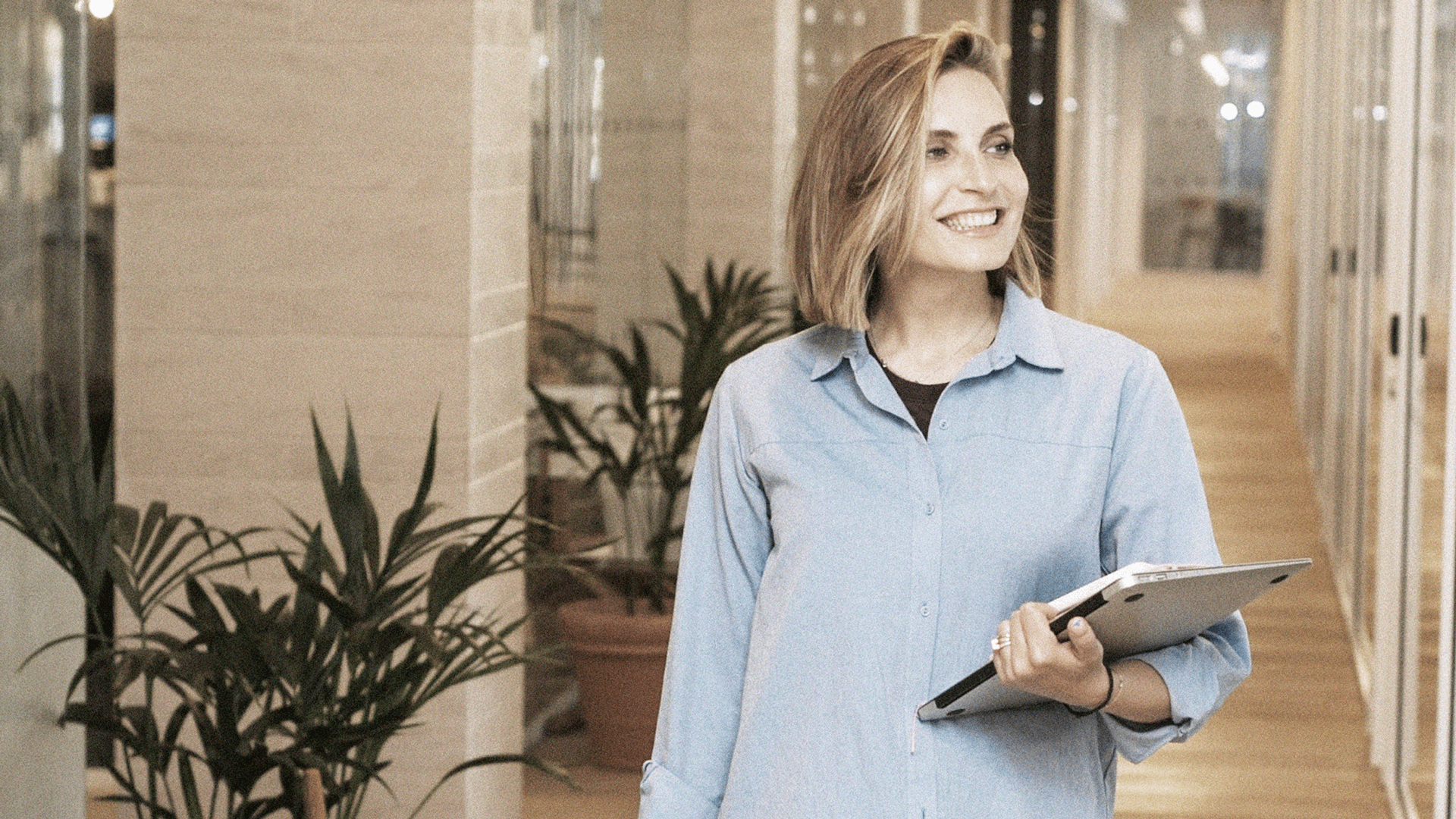 Eine lächelnde Frau in einem modernen Bürogebäude, die einen Laptop unter dem Arm trägt. Sie steht in einem hellen Flur mit Glaswänden und Topfpflanzen, was eine professionelle und einladende Arbeitsumgebung vermittelt.